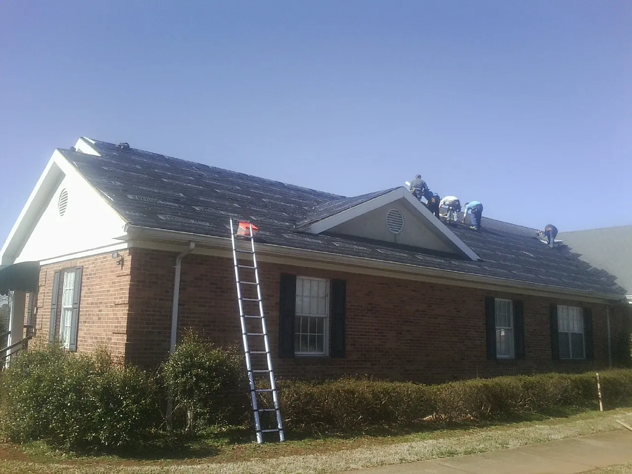 Crew tearing shingles off a brick ranch roof with a ladder set — Emergency Roof Repair work by our crew in Helenwood