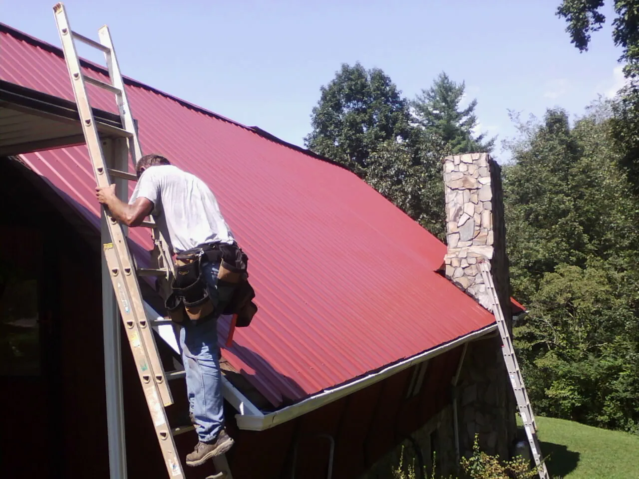Roofer on a ladder beside a red metal roof and stone chimney — Soffit & Fascia Repair by Roofing Brothers in Helenwood